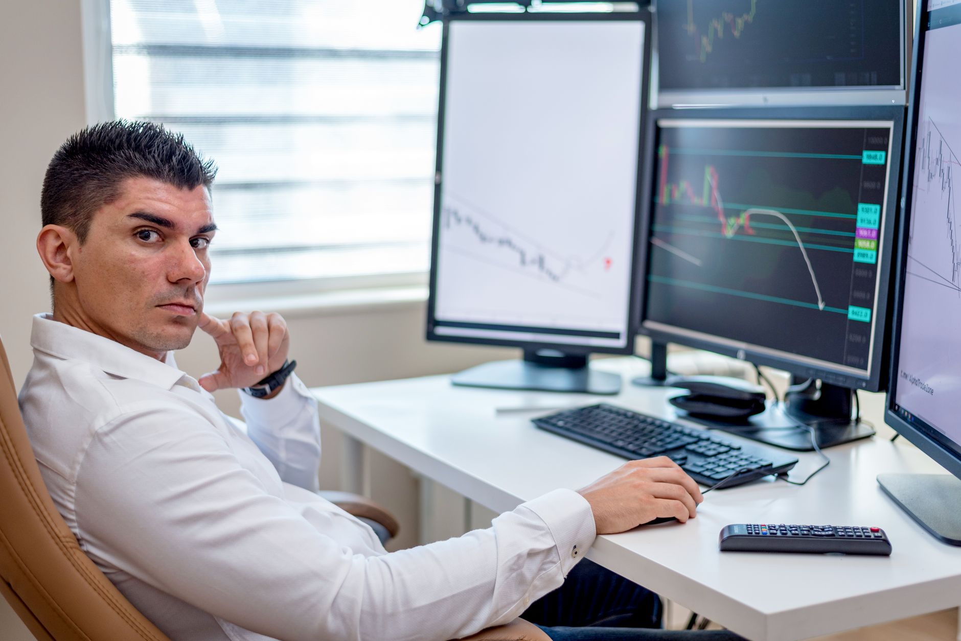 Homme en chemise blanche assis à un bureau devant plusieurs écrans affichant des graphiques financiers, le regard tourné vers l’objectif.