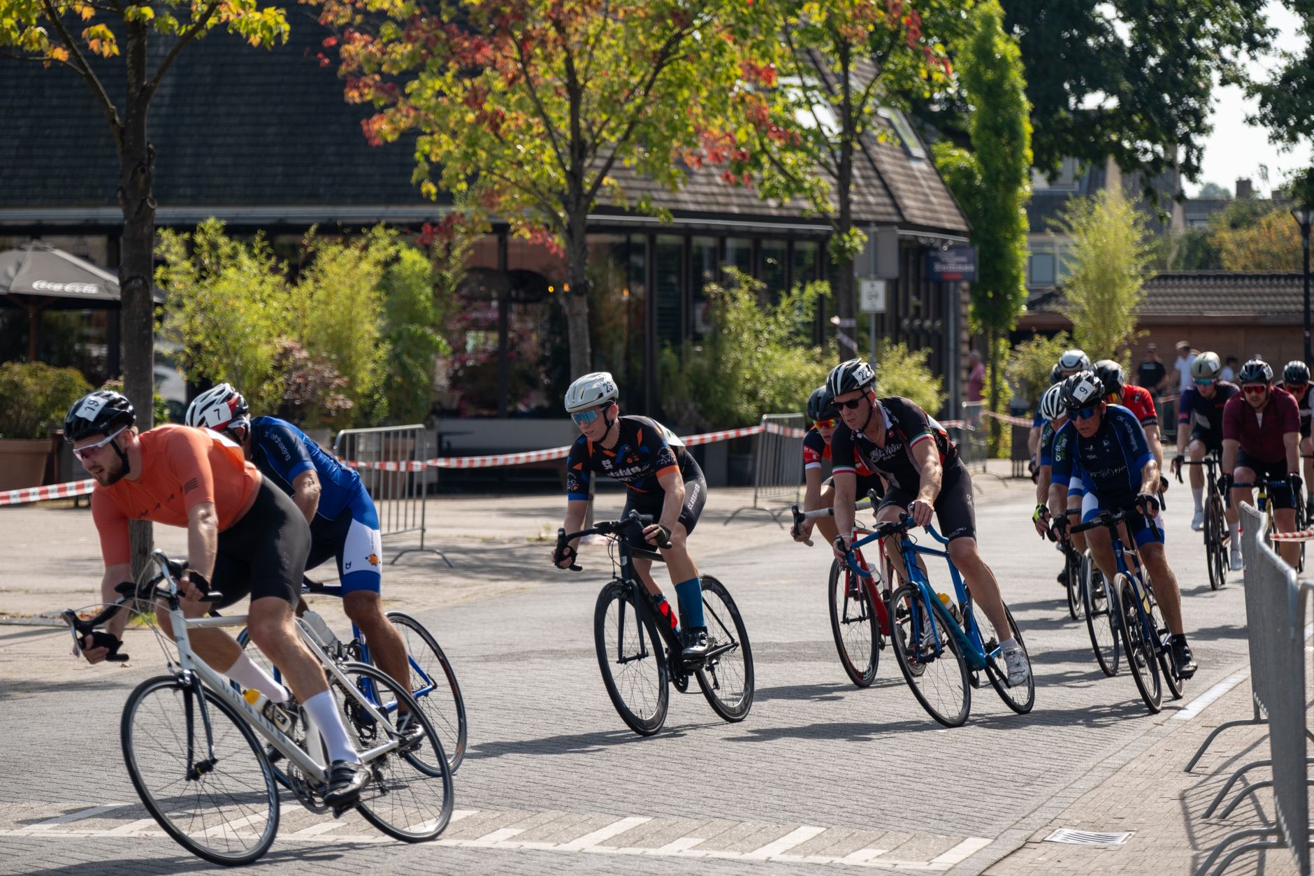Peloton de cyclistes en maillots colorés et casques roulant dans une rue pavée bordée de barrières, devant un bâtiment bas et des arbres.